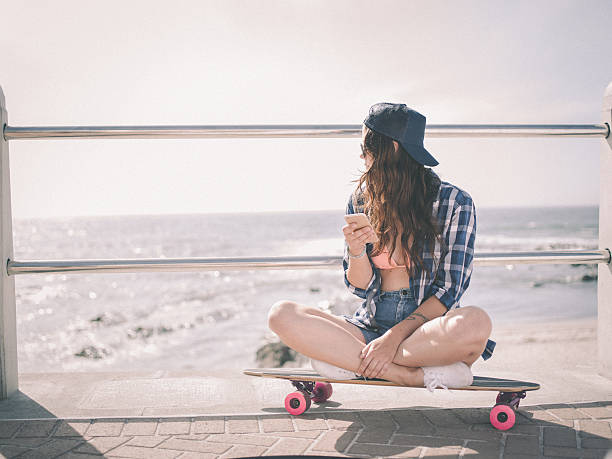 hipster de menina sentada em seu skate em frente à praia - teenager smart phone young women teenagers only - fotografias e filmes do acervo