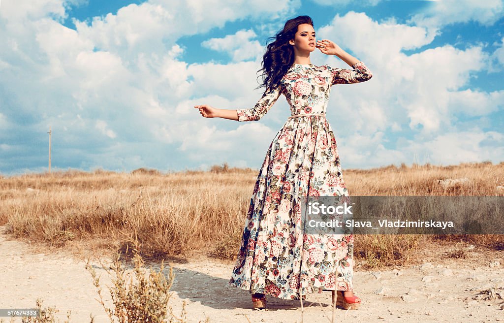 beautiful woman in luxurious floral dress posing in summer field fashion outdoor photo of beautiful woman with dark curly hair in luxurious floral dress posing in summer field Adult Stock Photo