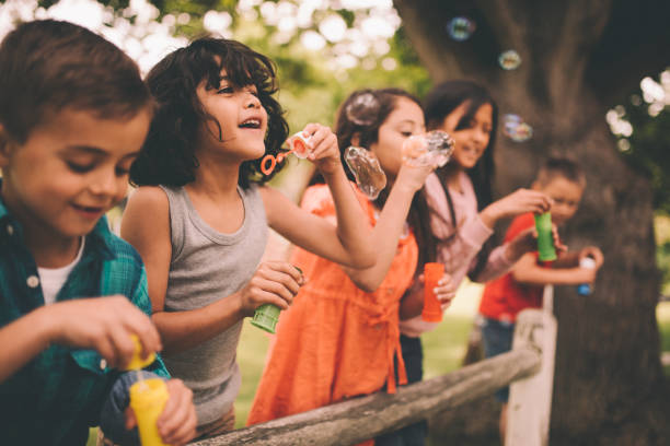 Little boy having fun with friends in park blowing bubbles Long haired hispanic boy laughing and having fun with his friends standing on a wooden fence in a summer park blowing bubbles, with a vintage develop summer stock pictures, royalty-free photos & images