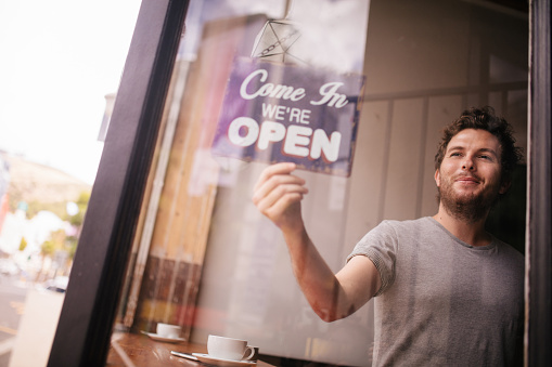 A smiling and positive hipster man turning an opening sign on glass front door of coffee shop with reflection of street in glass