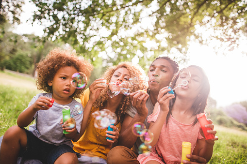 Young african-american family sitting and playing together in beautiful park blowing bubbles and laughing