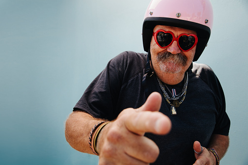 Silly senior adult man with pink safety helmet and red heart shape sunglasses, isolated on blue wall background