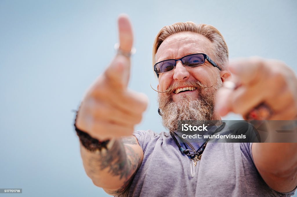 Smiling good-natured man looking into the camera Bearded mature man pointing and looking at camera, isolated on blue wall background Men Stock Photo