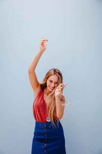 Beautiful teenage girl with long blond hair wearing trendy boho outfit and dancing, isolated on blue wall background