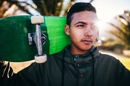 Portrait of mixed race person holding a skateboard behind his head, outside, with the sun behind him