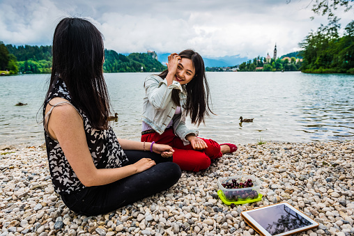 Two beautiful young Chinese women with dark long hair sitting on the pebbles on the Bled lake shore and eating cherries. One in red trousers is laughing. The other in black is turned towards the lake(rear view). Digital tablet and the cherry box on the floor. The castle and the island with a church in the background. Cloudy summer day, Bled, Slovenia, Europe. Nikon D800, full frame, XXXL.