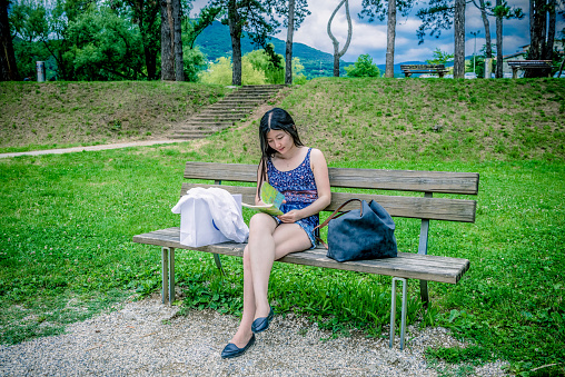 A pretty young Chinese woman with dark long hair, wearing blue singlet and shorts, is sitting on a park bench and reading from her notebook. Front view. She has crossed her legs. Beside her dark handbag and white shopping bag. Trees and grass in the background, summer,  Europe. Nikon D800, full frame, XXXL.  Copy space.
