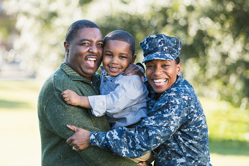 A military homecoming. Portrait of an African American woman wearing navy camouflage uniform standing outdoors with her family. Her husband is holding their 5 year old son who is in the middle between his parents, smiling at the camera. The main focus is on the woman.