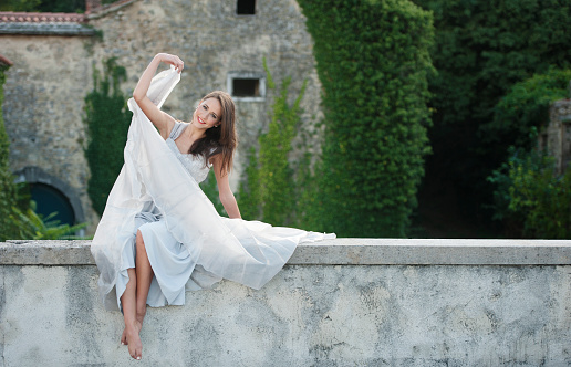 Beautiful young female fashionmodel in white silk evening dress sitting on a wall of an old house. Slovenia.