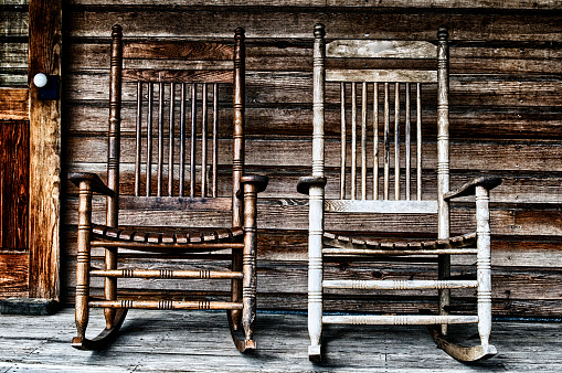 Two old wooden rocking chairs on front porch, part of door and the house's wooden shingles can be seen.