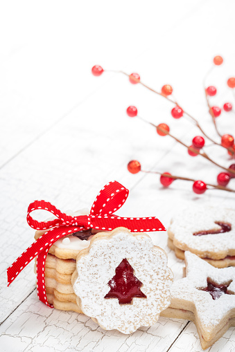 Christmas tree linzer cookies  decorated with powdered sugar and with red jam center over a white background. Red bow and berries finish this pretty, high-key, border.