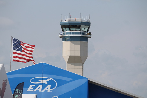 Red and white colored airport radar tower with many steps going up to the top of the antenna and the front captain's cabin of a white aircraft. Dramatic cloudy sky and air travel background, copy space.