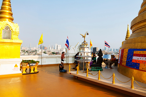 Bangkok, Thailand - February 19, 2013: Capture of some praying Thai women on roof of Wat Traimit, socaled golden mountain in afternoon sunshine, At wall are some thai flags. At right side is golden pagoda. At left side is small pagoda and exit of roof.