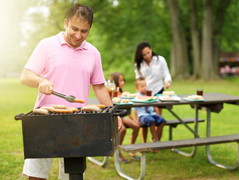 father grilling hot dogs and bratwurst for family at park