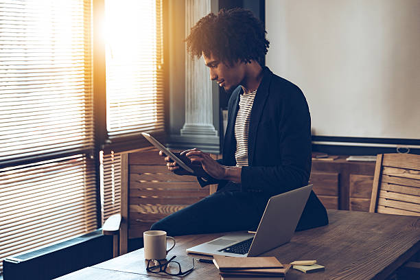 Managing his timetable. Side view of young handsome African man using his digital tablet while sitting on table at his working place stubble male african ethnicity facial hair stock pictures, royalty-free photos & images