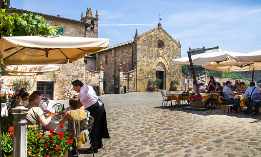 Monteriggioni, Italy, May 30, 2015 - waiter serving food on tourist restaurant in the main square with the Santa Maria Assunta church on background
