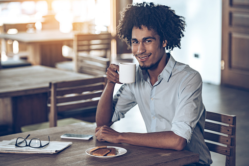 Young African man holding coffee cup and looking at camera with smile while sitting in cafe