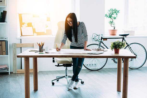 Working on new project. Full length of young beautiful Asian woman making some notes at blueprints while leaning to the table in office legs crossed at ankle stock pictures, royalty-free photos & images
