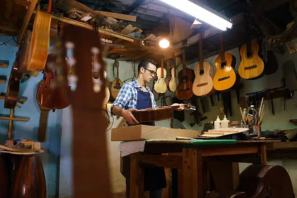 Lute maker shop and acoustic music instruments: a young adult artisan fixes an old classic guitar, then stores it in a cardboard case for his client. Wide shot