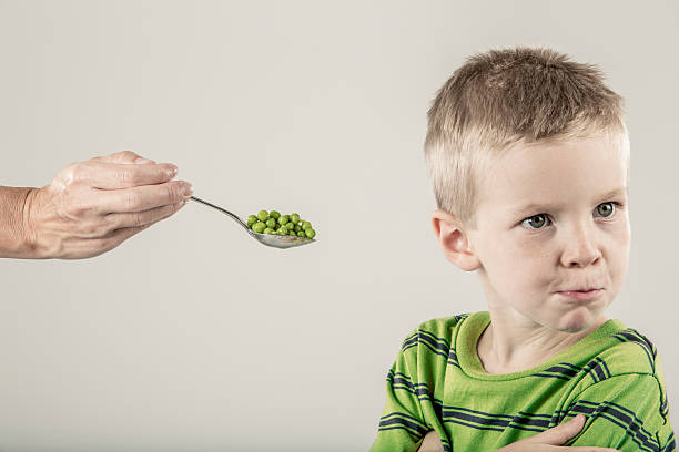Young Boy Refusing a Spoonful of Peas A parent is attempting to feed a boy a spoon piled high with green peas. The boy is turning his head and refusing to eat the vegetables. The boy's is wearing a green shirt that matches the color of the pile of peas. The background is a neutral off-white (stock image) happy green peas stock pictures, royalty-free photos & images
