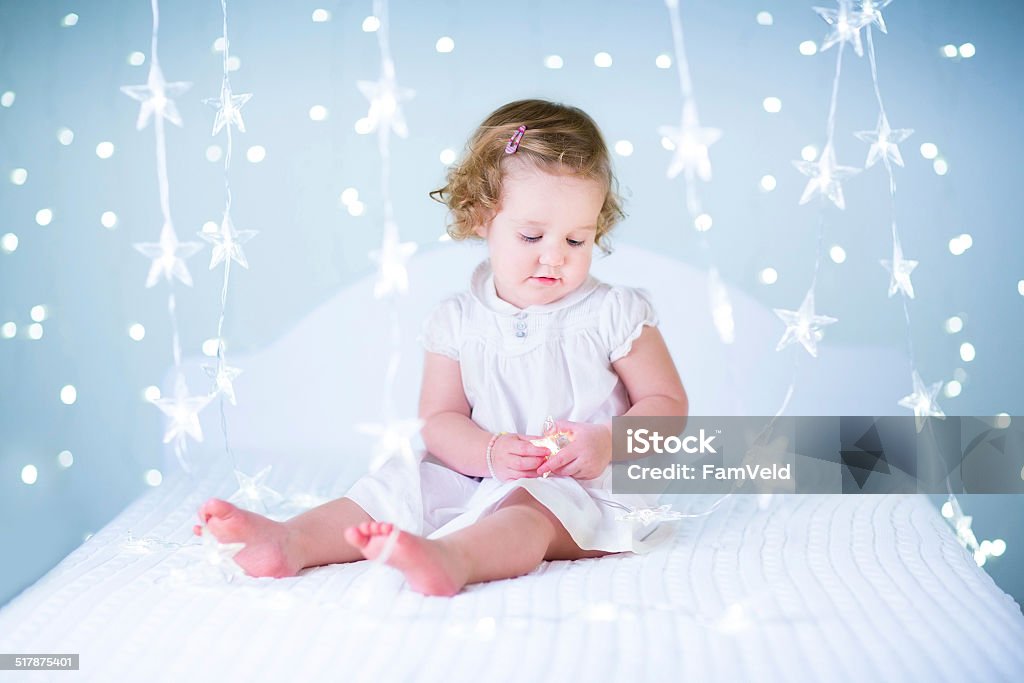 Adorable toddler girl playing with Christmas lights on white bed Adorable toddler girl playing with Christmas lights sitting on a white bed Child Stock Photo