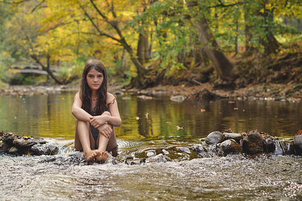 a girl in a bathing suit sits in a shallow river or stream and looks directly at the camera.  The trees in the background are yellow, it is fall in New England, they are reflected in the water.  She is very calm, barefoot with long hair. 