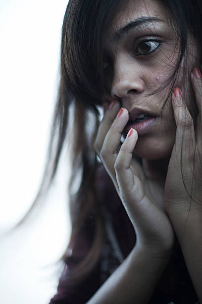 Frightened girl holding her face and giving intense expression. Indoor image of a late teen Asian/Indian girl holding her face and giving fearful intense expression by her eyes and mouth remained open in a shock. One person, vertical composition with selective focus and copy space. domestic violence india stock pictures, royalty-free photos & images