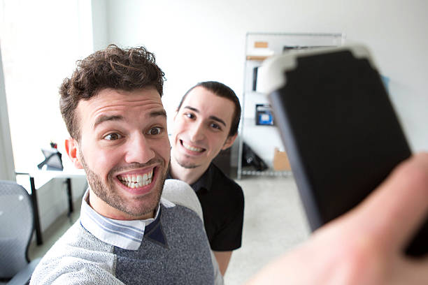 Young Businessmen Taking Selfies in an Office Two young businessmen make silly faces while taking selfies in a modern office. photo bomb stock pictures, royalty-free photos & images