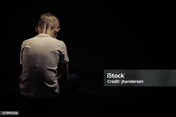 Rear View Of A Seated Sad Young Boy Against Black Stock Photo - Download Image Now - Child, Sadness, Dark