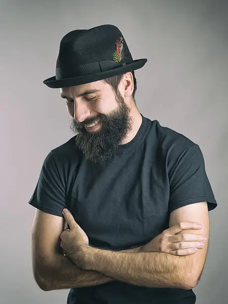 Bearded hipster wearing black t-shirt and hat laughing spontaneous with closed eyes. Retro toned filtered portrait over gray background with vignette effect