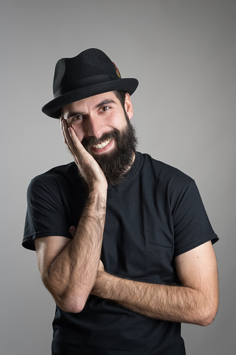 Friendly smiling bearded hipster wearing black t-shirt and hat with head resting on his hand. Headshot portrait over gray studio background with vignette