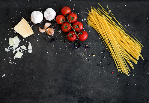 Food frame. Pasta ingredients. Cherry-tomatoes, spaghetti pasta, garlic, basil, parmesan and spices on dark grunge backdrop, copy space, top view, horizontal oriented
