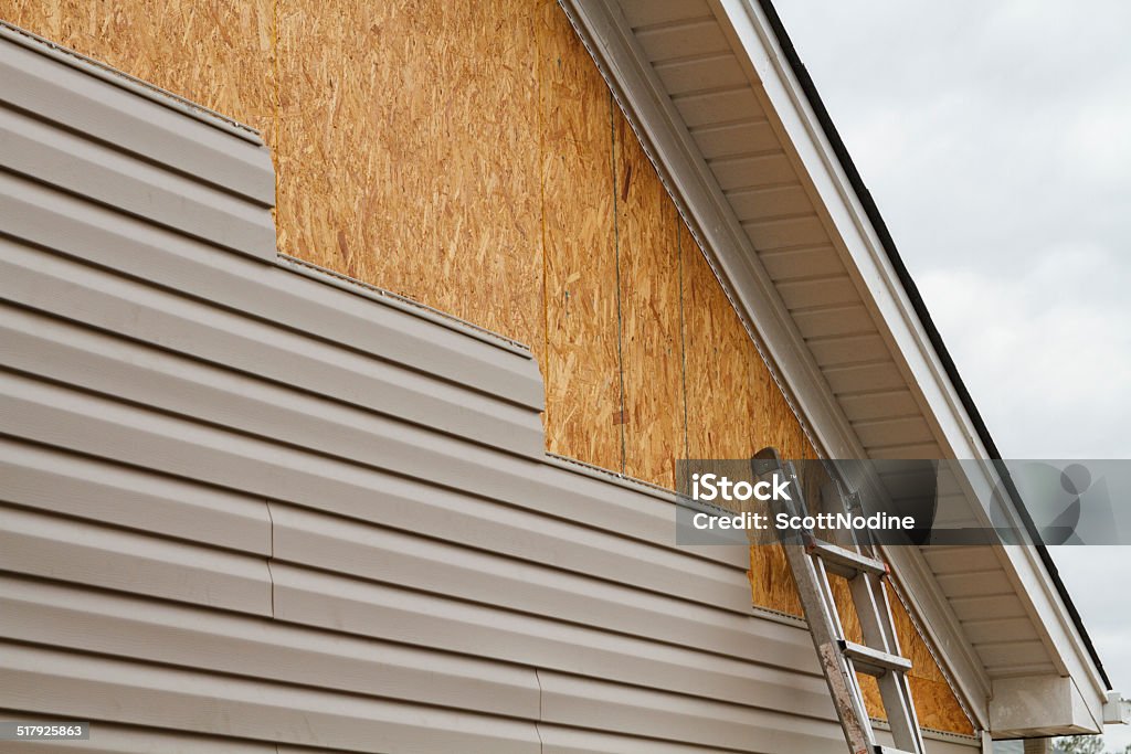 Vinyl Siding Installation On A House In The South New beige vinyl siding being installed over an osb (oriented strand board) substrate on a residential house in the Southeastern USA region on a cloudy day. The old vinyl siding was being replaced after a hail storm. Siding - Building Feature Stock Photo