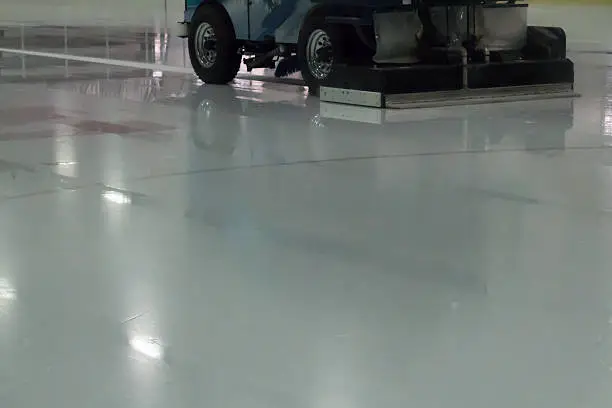 Ice wet and being resurfaced at a hockey rink.  The bottom of a ice resurfacer can be seen, with the wheels driving over the ice of the rink.  The machine scrapes the ice and lays down warm water to freeze.
