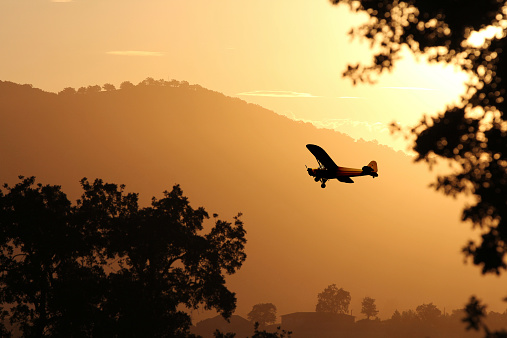 A small silhouette of a  Cessna airplane is flying in a beautiful sunset of orange and yellow through the mountains and trees.