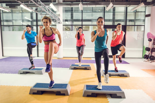 Group of athletic people exercising step aerobics in a gym. Medium group of smiling young people having step aerobics training in a health club. aerobics stock pictures, royalty-free photos & images