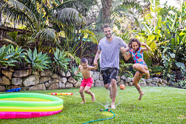 Father and children jumping in sprinkler Father and children jumping in sprinkler in the backyard garden. agricultural sprinkler stock pictures, royalty-free photos & images