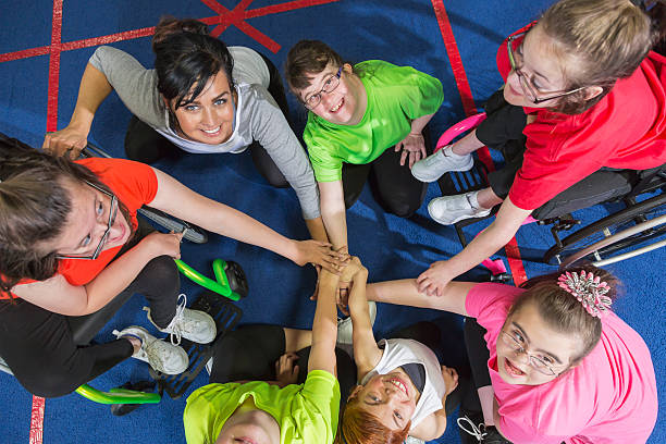 Group of special needs girls showing team spirit Overhead view of a group of special needs teenagers and young women with instructor, showing team spirit. They are sitting in a circle on a gym floor, hands in the center. Several of the girls have downs syndrome and two are in wheelchairs. developmental disability stock pictures, royalty-free photos & images