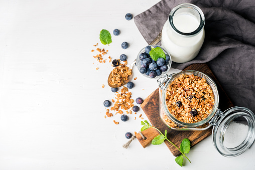 Healthy breakfast ingrediens. Homemade granola in open glass jar, milk or yogurt bottle, blueberries and mint on white wooden background, top view, copy space