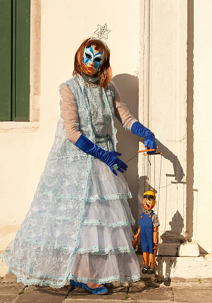 Venice Carnival 2014 Venice, Italy - February 24, 2014: The photo was taken in front the wall with green window on Campo San Zaccaria in Venice. Female puppeteer holding wooden puppet in fer hands and playing with it. She wearing red wig, blue gloves and white and blue silk carnival costume. Vertical color image. mardi gras body painting stock pictures, royalty-free photos & images