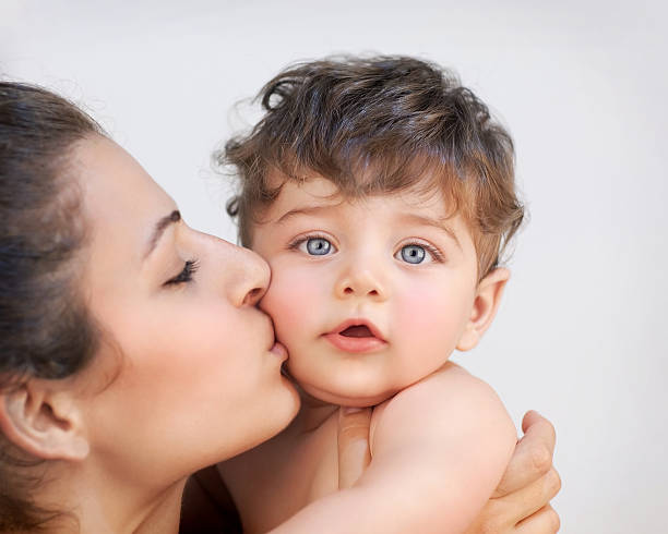 Closeup portrait of cheerful young mother kissing in cheek her precious little son, isolated on gray background, happy family life