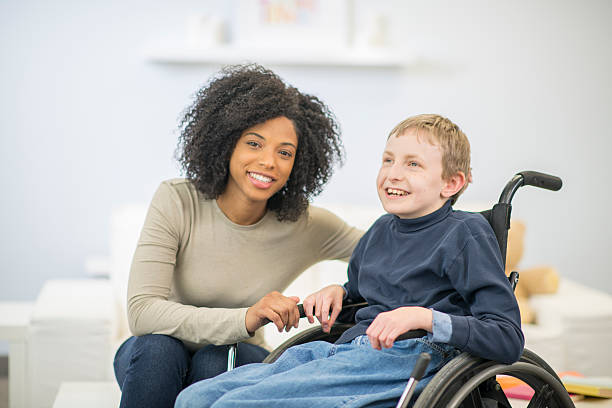 Young Boy in a Wheelchair A nurse or caregiver is helping a boy with cerebral palsy get into his wheelchair. They are both smiling happily together. special education stock pictures, royalty-free photos & images