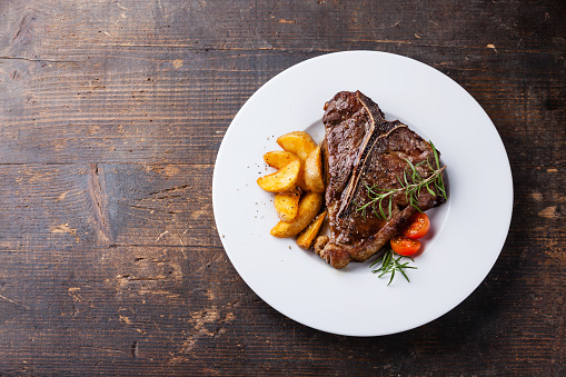 T-Bone Steak with roasted potato wedges on white plate on wooden background