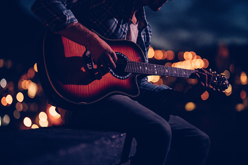 Close up of a teenage man playing an acoustic guitar on a rooftop of  a building at night with citylight in the background