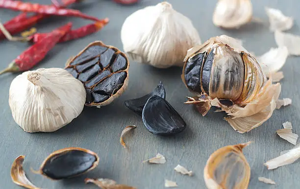 Black garlic, whole, in cloves and a cross-section, scattered on grey back ground with chili peppers