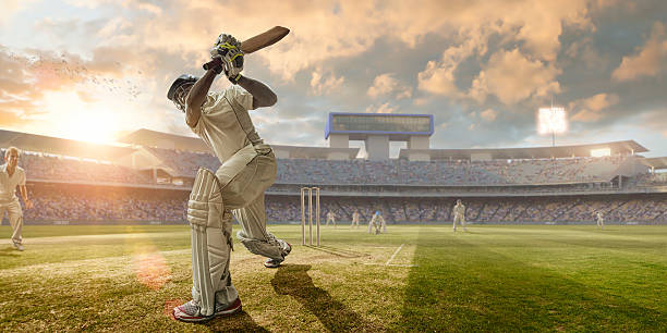 Cricket Batsman Hitting Ball During Cricket Match In Stadium A close up image of a professional cricketer playing in batsman position wearing cricket whites and safety helmet, having just hit a ball during a cricket match in an outdoor stadium full of spectators. The action occurs under an evening sky at sunset. Stadium is fake, created from photographic and CG elements.  cricket stock pictures, royalty-free photos & images