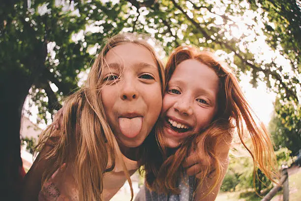 Two young girls who are best friends posing for the camera while one is laughing and the other is sticking her tongue out under a huge tree with green leaves in a sunny park