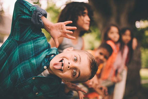 Little boy pulling a funny face with friends in background Vintage style shot of a cute little boy leaning over and pulling a silly face for the camera with friends standing on a fence smiling in the background in a summer park gurning stock pictures, royalty-free photos & images