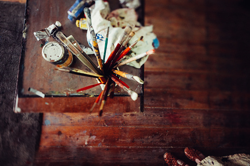 Topview of a wooden table in an art studio with a collection of paintbrushes in a container and tins and tubes of paint alongside a rag
