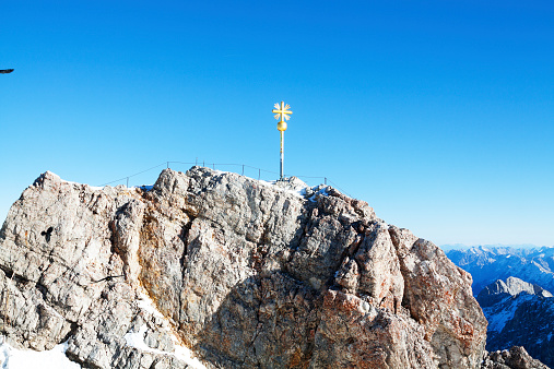 Golden cross of Zugspitze in blue and sinny sky, some birds are flying around.